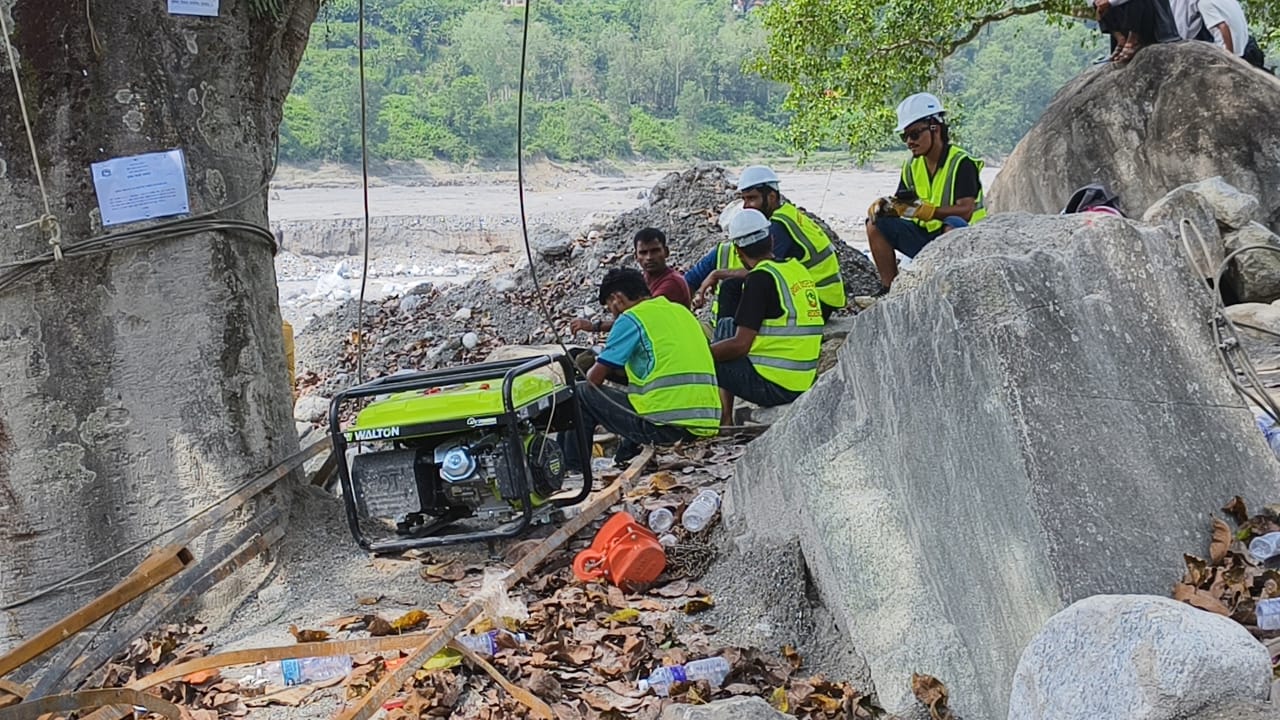 Workers in high-visibility vests and helmets with a generator among boulders by the river